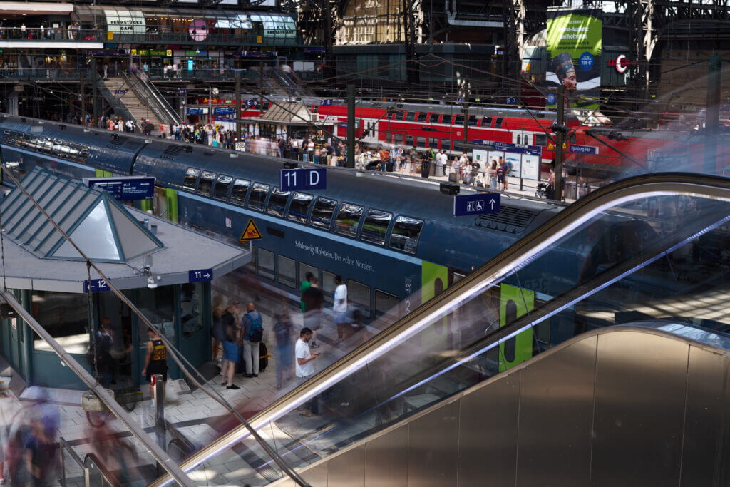 die Menschen auf der Rolltreppe nach oben sind nur als Schleier zu erkennen. Auf dem Bahnstteig stehen wartende Menschen, auf den hinteren Gleisen Züge